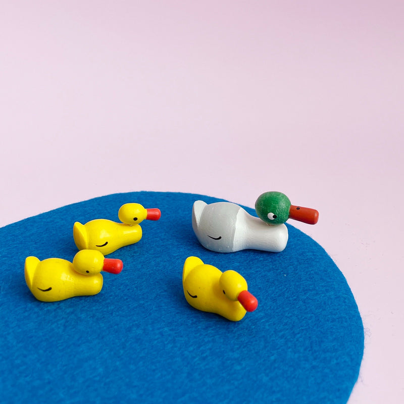 A small wooden duck figurine with three ducklings. They sit on a blue piece of felt on a pink background. 
