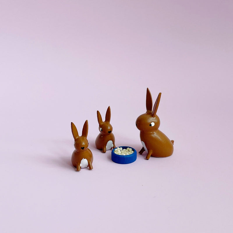 Three small  brown wooden bunny figurines on a light pink background. 