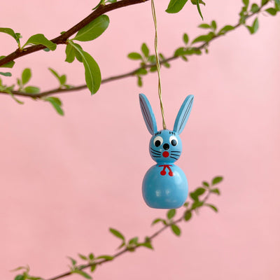 A light blue bunny ornament hanging on a branch on a pink background.