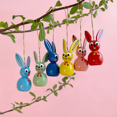 Six handpainted bunny ornaments hanging on a branch on a pink background. 