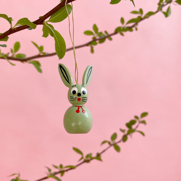 A light green bunny ornament hanging on a branch on a pink background.