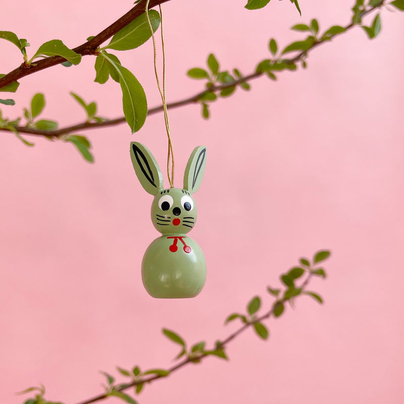A light green bunny ornament hanging on a branch on a pink background.