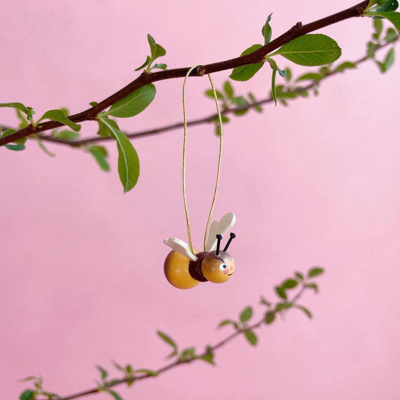 Wooden bee ornament hanging from a branch on a pink background.