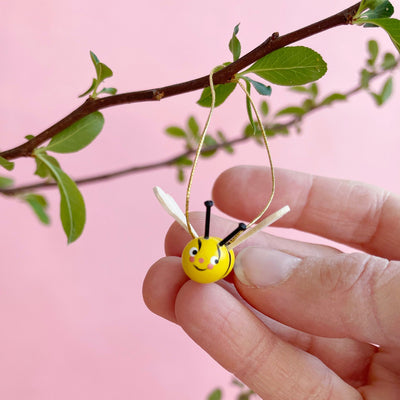 Wooden bee ornament hanging from a branch, and being held by fingertips,  on a pink background.