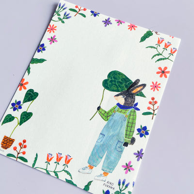 The writing paper of the letter set features a grey and beige anthropomorphic rabbit wearing a green shirt and overalls. The rabbit holds a large leaf. The paper is bordered with bright-colored flowers and plants.