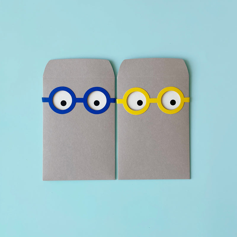 Two gray envelopes laying next to each other. The envelopes have eye holes cut out and you can see white cards with eye dots printed on them peeking through.  The left envelope has blue glasses around the eyes the left envelope has yellow glasses. 