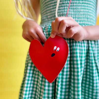 Red heart shaped purse being worn and held by a child in a green gingham dress