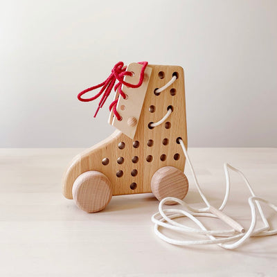 Wooden roller skate shaped lacing toy with red and white laces on a cream background.