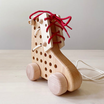 Wooden roller skate shaped lacing toy with red and white laces on a cream background