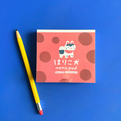 a small note pad illustrated with red polkadots and a cat on a bright blue background. A yellow pencil lies on the left. 