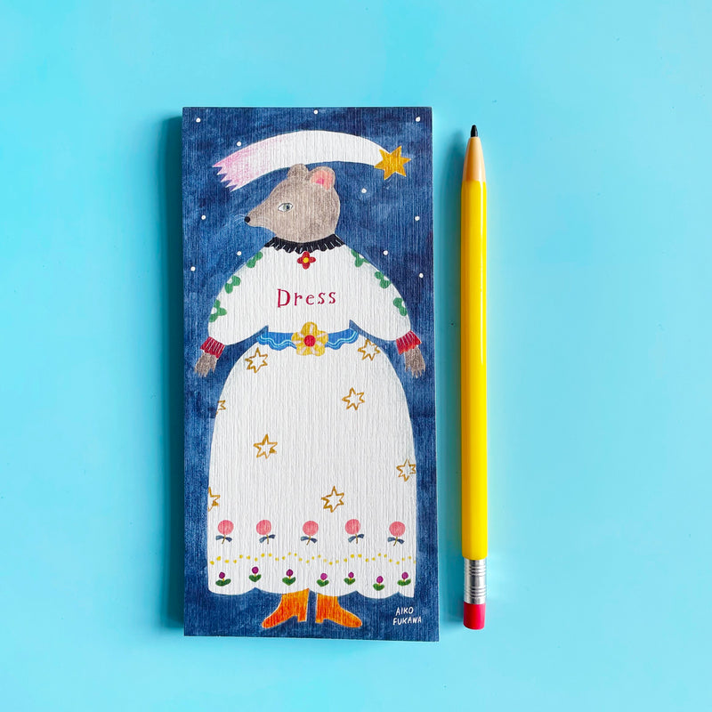 The cover of the Aiko Fukawa Dress Memo Pad on a blue background, next to a pencil for scale. The cover shows a grey anthropomorphic mouse standing in a white dress under a shooting star.