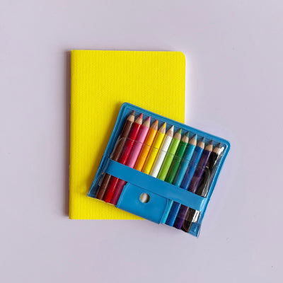 Tiny set of colored pencils in a blue package sitting on a small yellow notebook against a light purple background. 