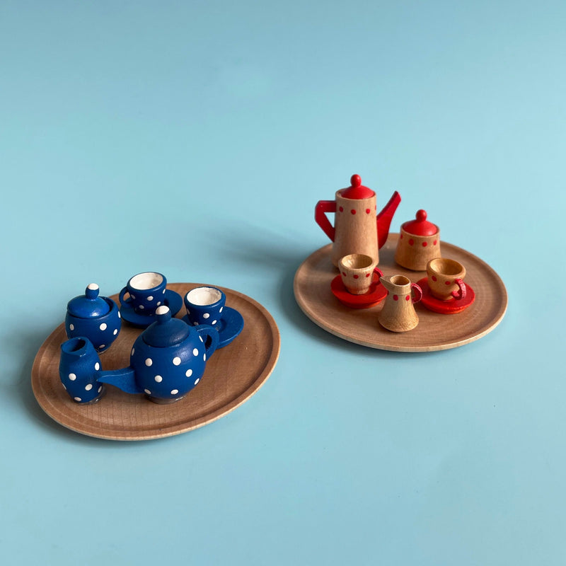 Two wooden tea sets on a light blue background. The tea set on the left is blue with white polka dots. The one on the right is red and natural wood colored. 